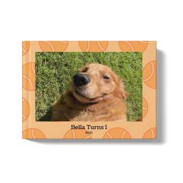 Pets Photobook with an orange background and tennis ball pattern. At the center of the cover is an image of a golden retriever lying in the grass, with a title beneath the image that says: "Bella Turns 1, 2025"